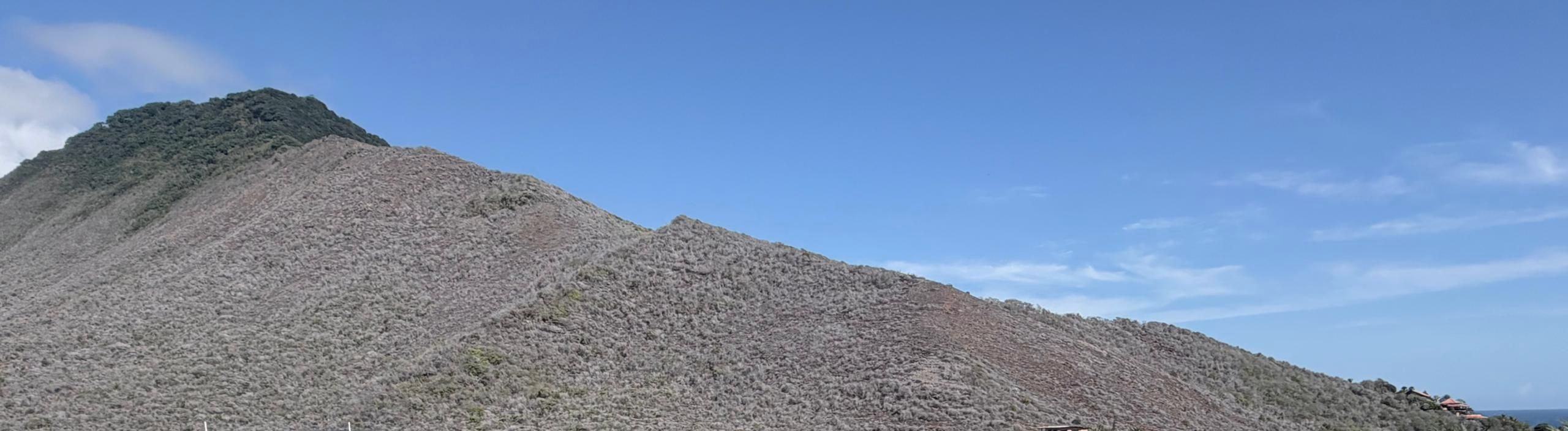El cerro detrás de Los Geckos bajo un cielo despejado, con el mar asomando en el borde derecho. Los Geckos · foto de obra · octubre 2026.