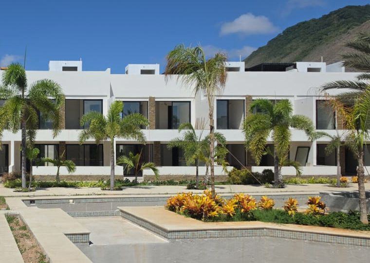 Hilera de módulos de Los Geckos con palmas y crotos, el cerro al fondo y una piscina vacía en primer plano. Los Geckos · foto de obra · octubre 2026.