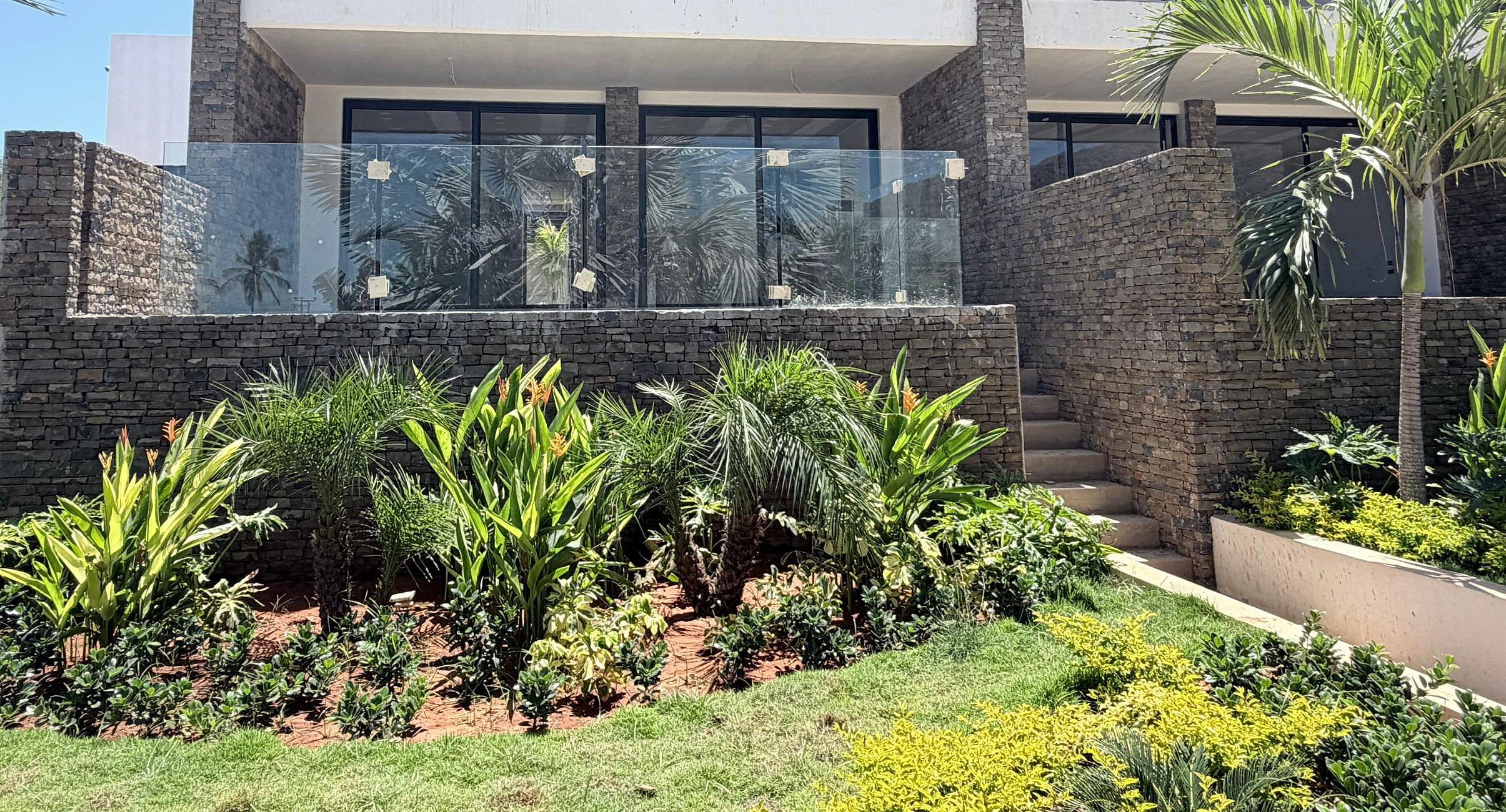 Fachada de Los Geckos con muro de piedra seca, baranda de vidrio y jardín tropical al frente. Los Geckos · foto de obra · octubre 2026.
