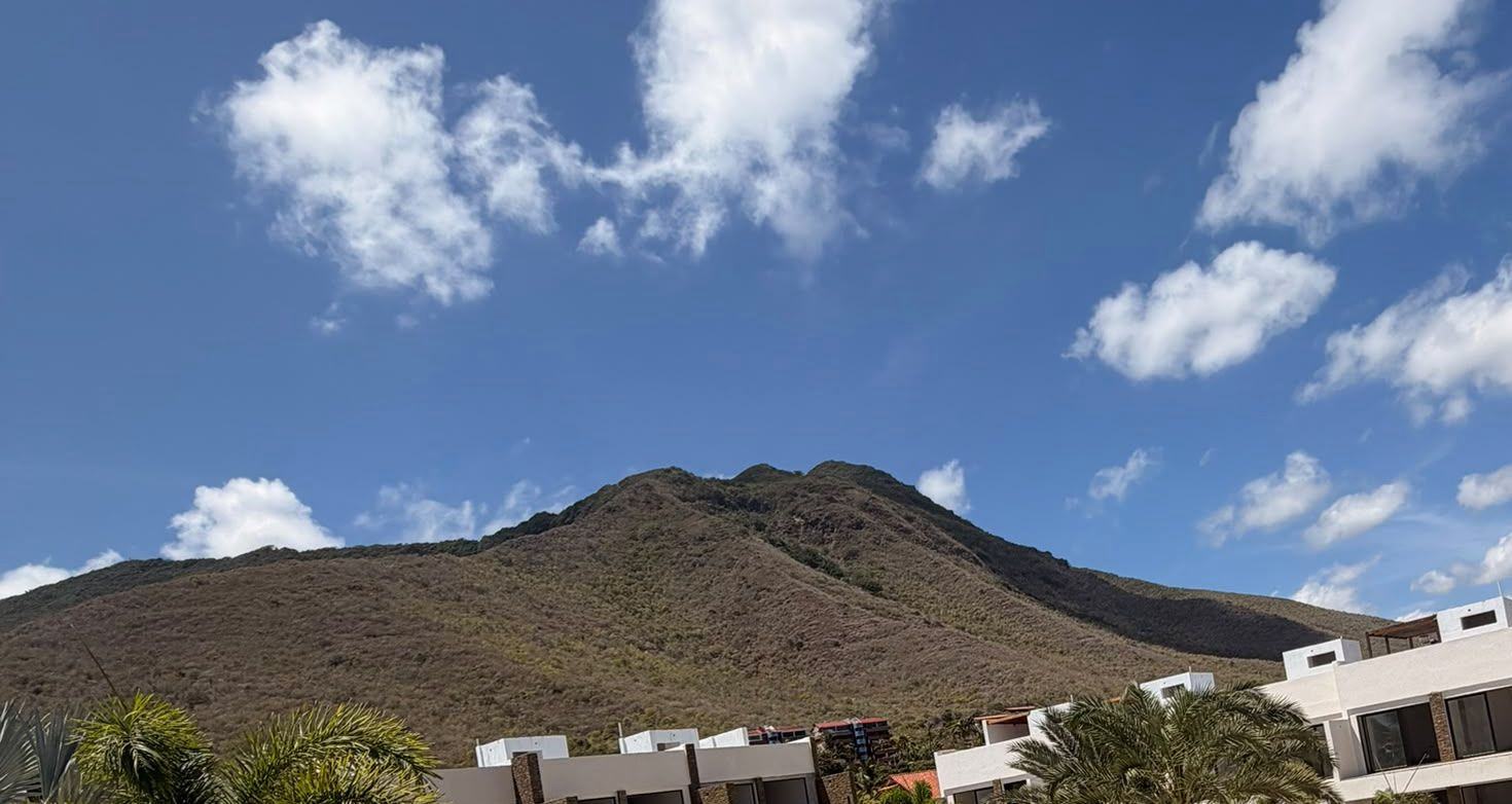 El cerro bajo un cielo con nubes, con los módulos blancos de Los Geckos y palmas en la parte baja. Foto de obra.